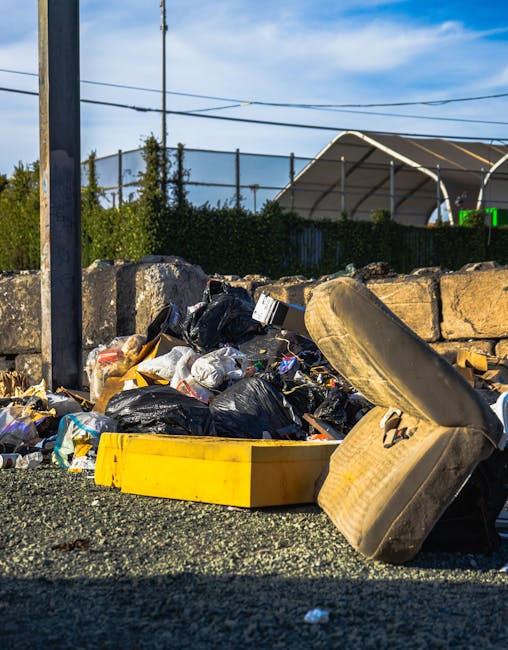 A person wearing white sneakers, black shorts, and a light blue t-shirt is using a grabber tool to pick up a piece of litter from a rocky area, which appears to be part of a shoreline or riverbank wit